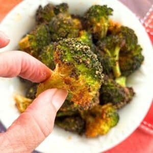 Hand holding a crispy Parmesan broccoli bite with golden, cheesy edges over a bowl of roasted broccoli.