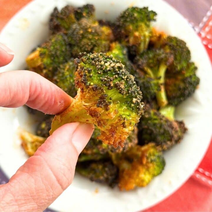 Hand holding a crispy Parmesan broccoli bite with golden, cheesy edges over a bowl of roasted broccoli.
