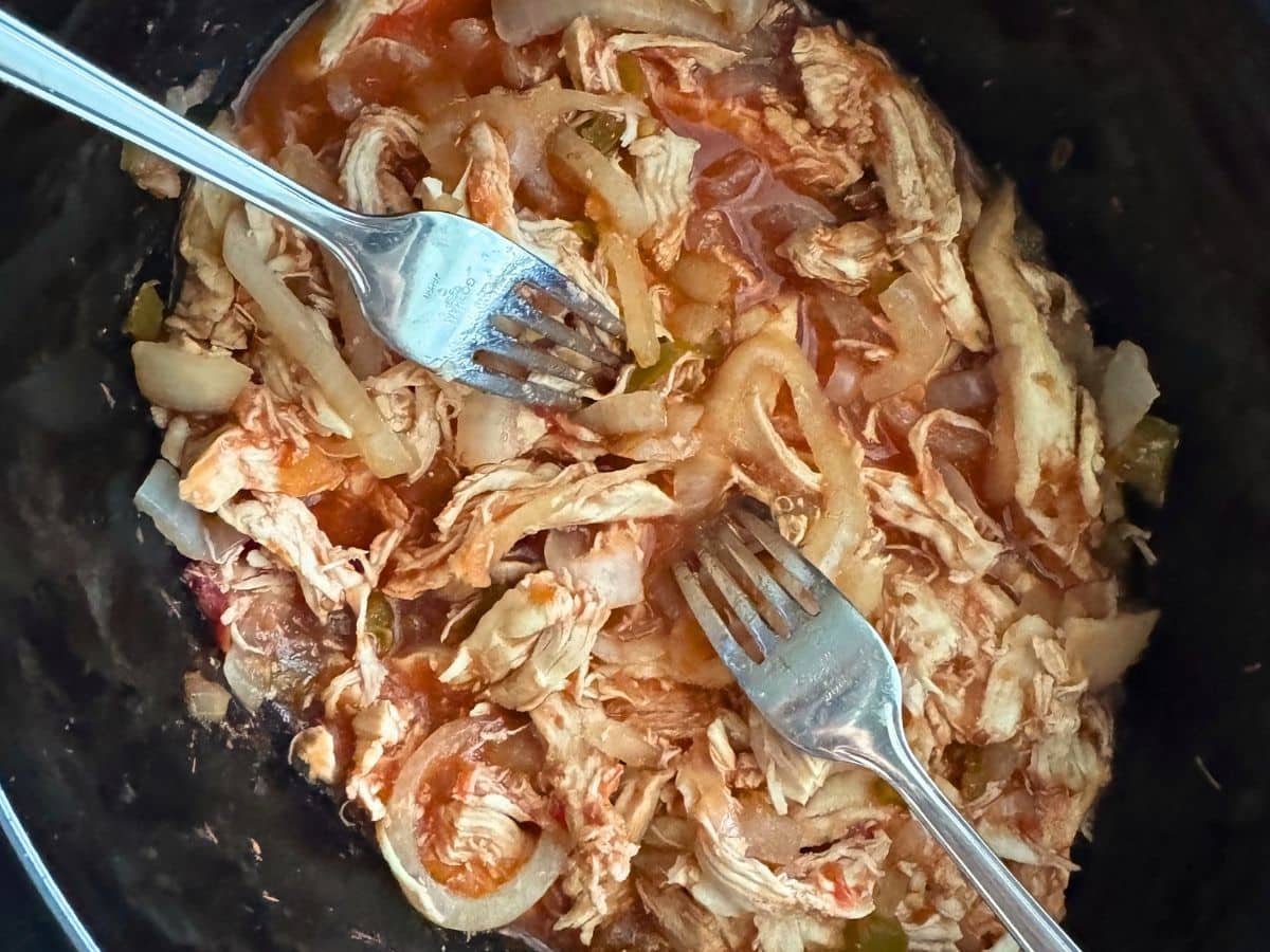 Shredding salsa chicken and cooked onions with two forks in a slow cooker.
