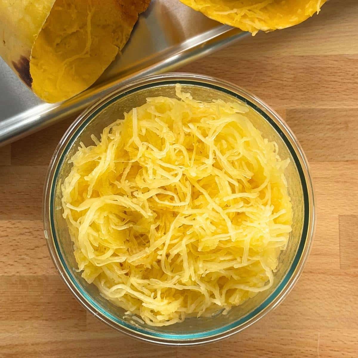 Spaghetti squash strands in a glass bowl in front of sheet pan and empty spaghetti squash gourds.