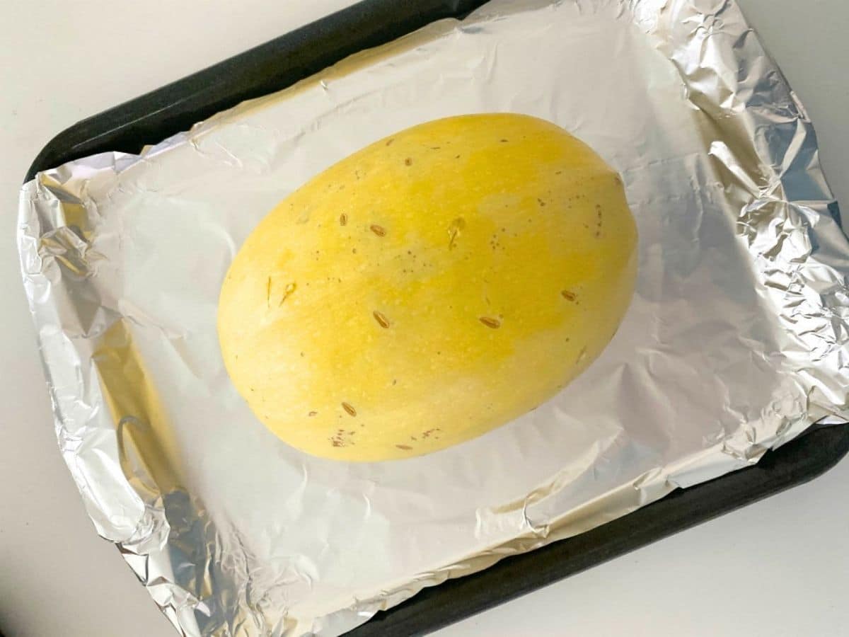 A whole spaghetti squash with knife piercing marks on a sheet pan covered in aluminum foil.