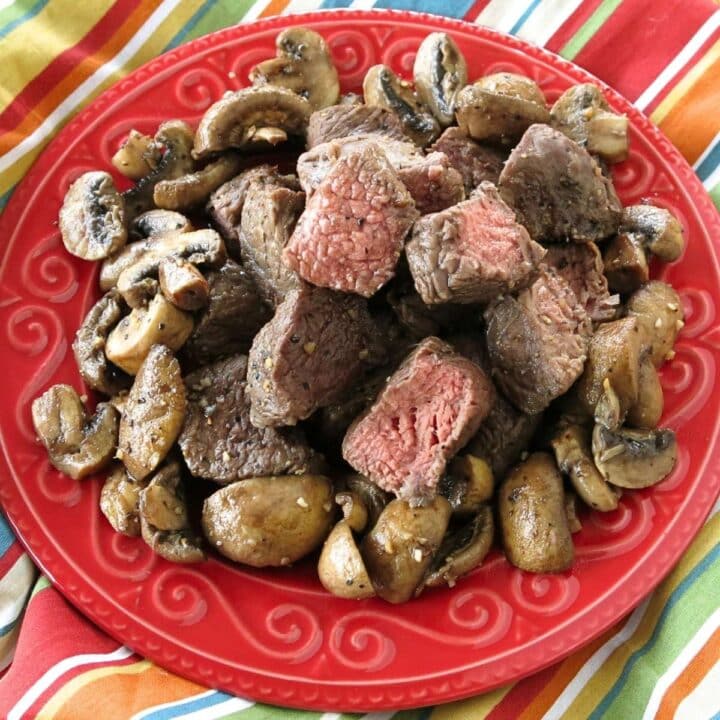 Juicy air fryer steak bites with browned edges and sautéed mushrooms piled on a red plate, with cut pieces showing pink centers.