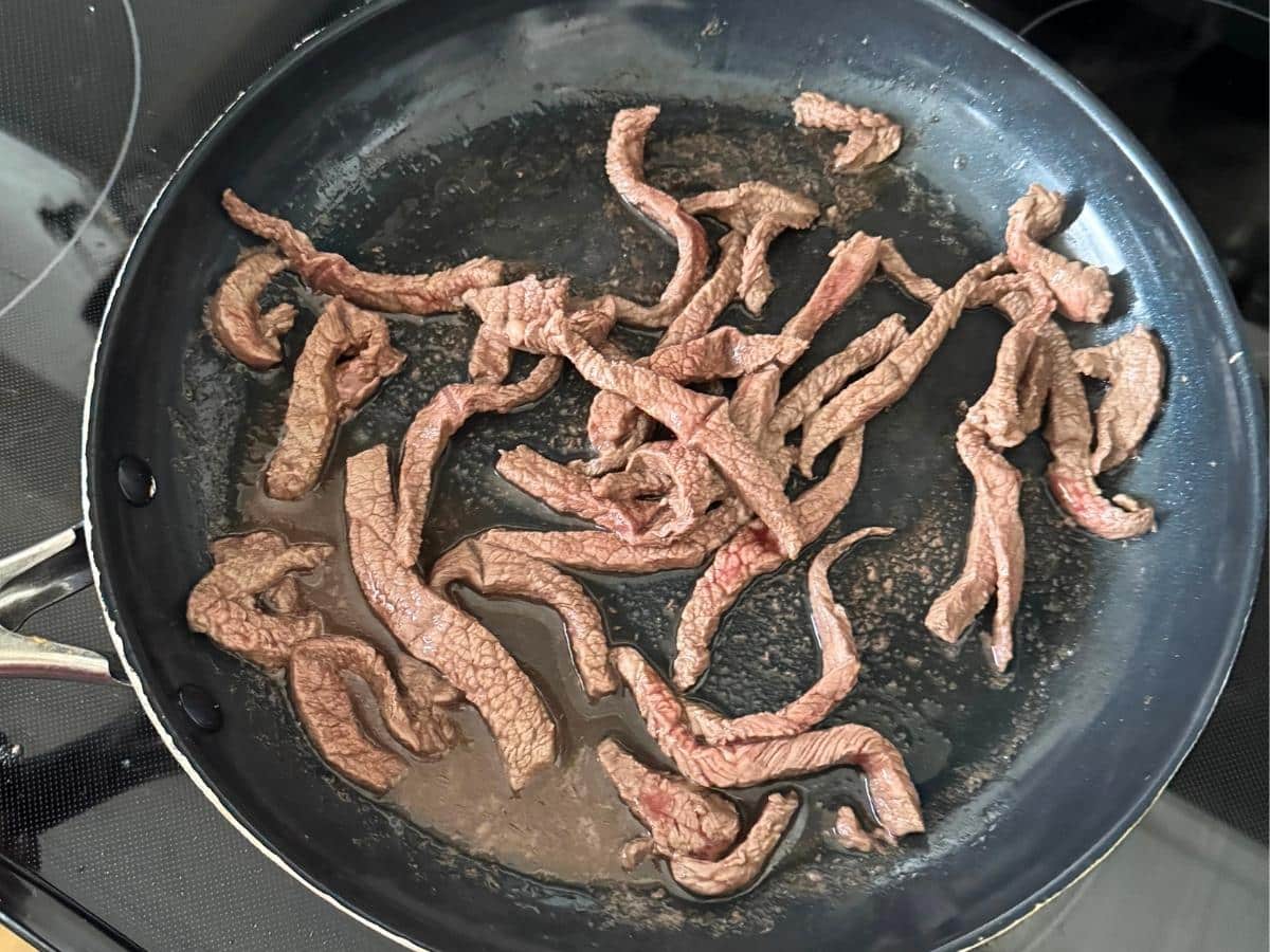Thin strips of beef browning in a large skillet on the stovetop.