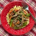Sesame beef stir fry with carrots, snow peas, green onions, and sesame seeds served over cauliflower rice on a red plate.