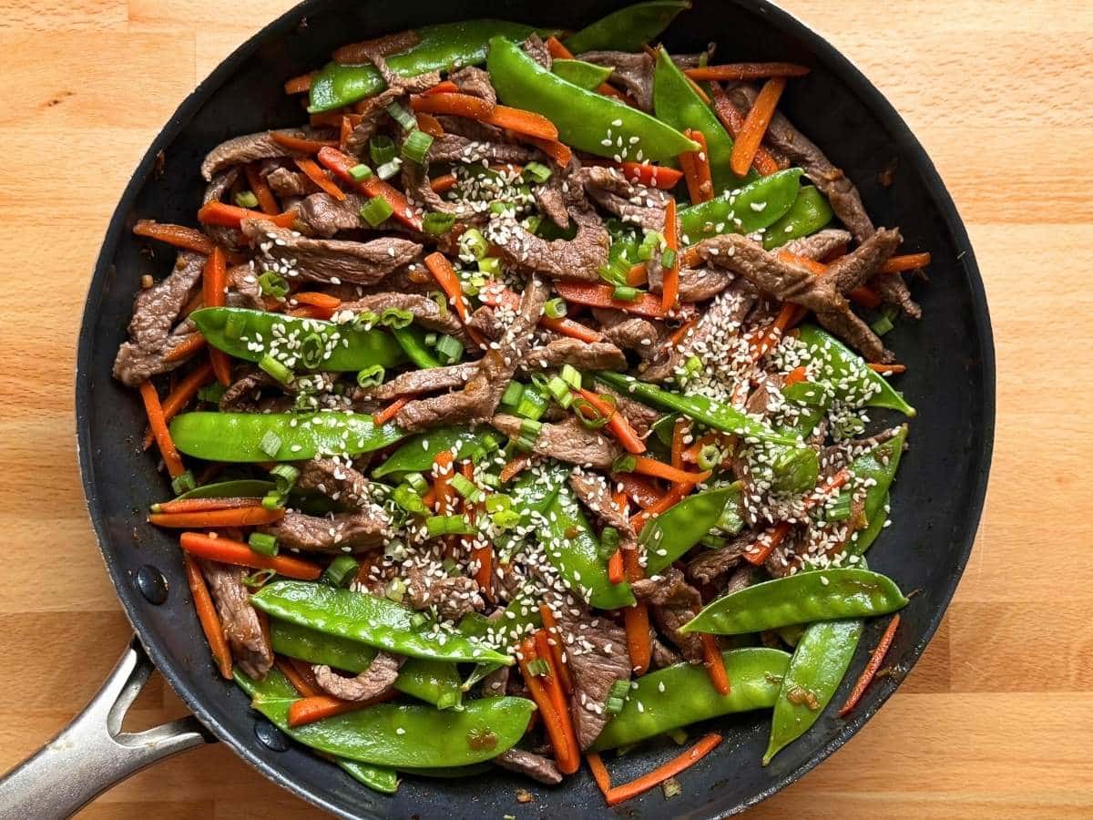 Sesame beef stir fry with thinly sliced beef, bright green snow peas, and carrots tossed together in a large skillet.