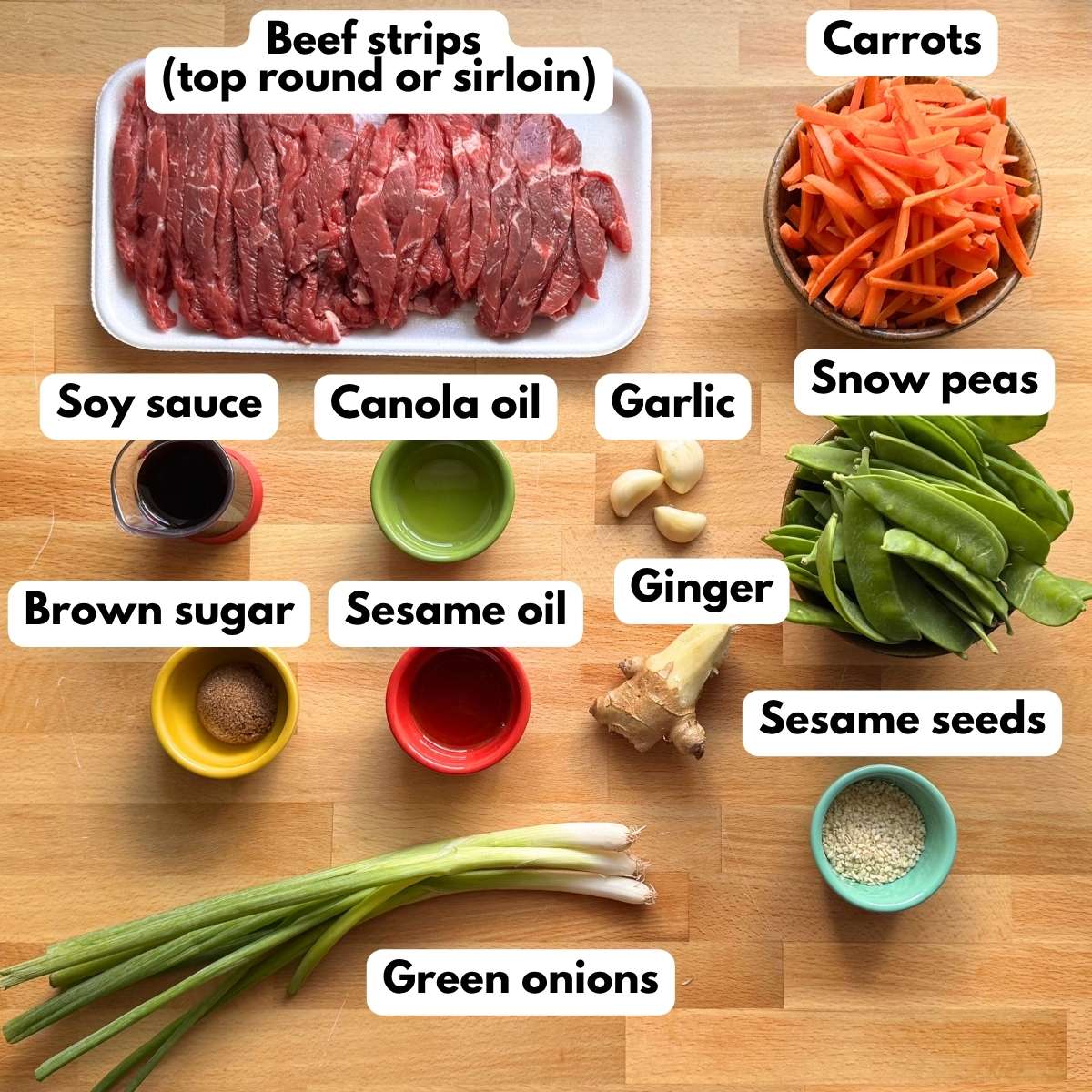 Ingredients for sesame beef stir fry, including beef strips, carrots, snow peas, fresh ginger, garlic, soy sauce, oils, brown sugar, green onions, and sesame seeds.