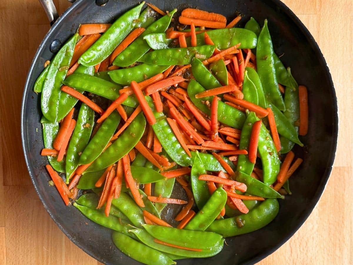 Snow peas and julienned carrots cooking in a skillet with ginger soy sauce until crisp-tender.