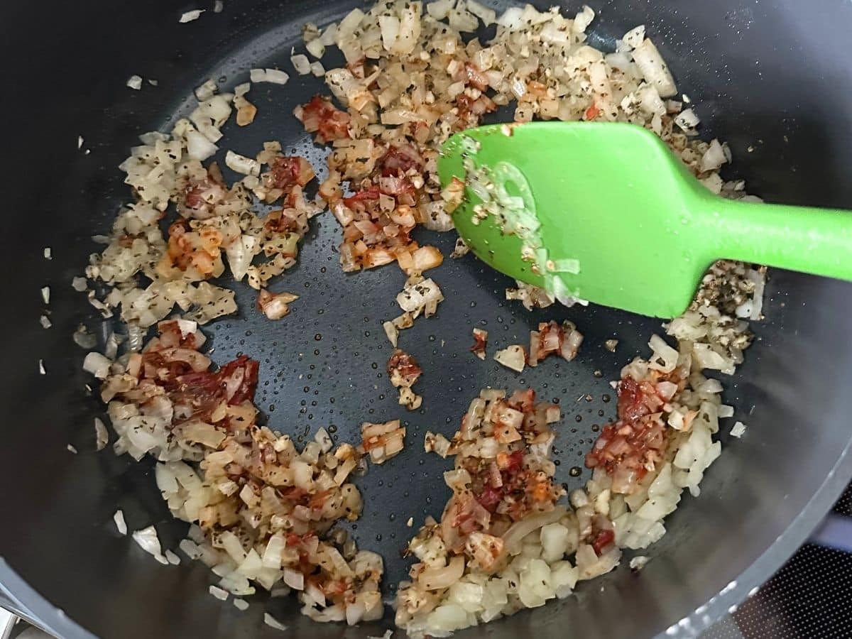 Diced onions, garlic, tomato paste, basil, and oregano sautéing in a large pot with a green spatula.